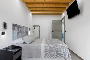 A bedroom with a bed, a TV mounted on the wall, a wooden ceiling, and a door leading to another room.