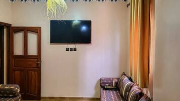 Ein Wohnzimmer mit einem an der Wand befestigten Flachbildfernseher, einem gemusterten Sofa, einer Holztür und einer Pendelleuchte.