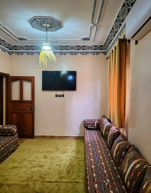 A living room with a flat-screen TV mounted on the wall, a patterned sofa, a wooden door, and a hanging light fixture.