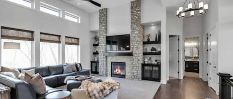 Amplia sala de estar con chimenea, ventilador de techo y vistas al exterior a través de grandes ventanales.