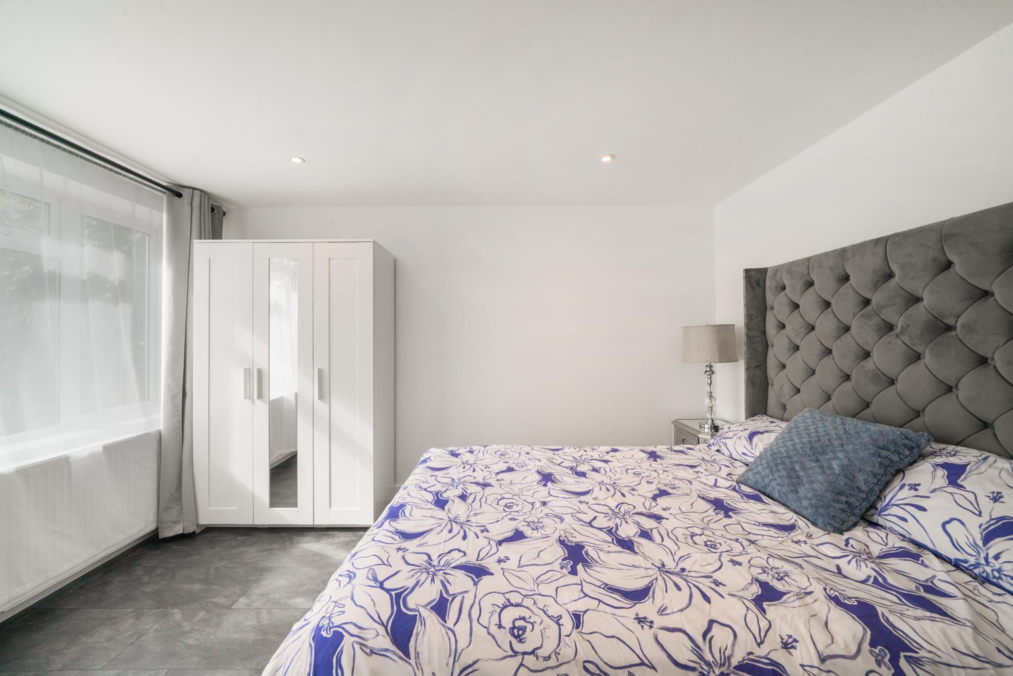 A bedroom with a large bed, a wardrobe, a bedside table with a lamp, and a view through a window.