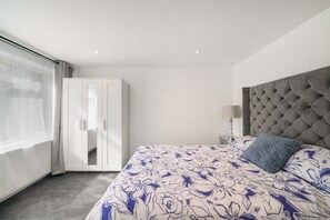 A bedroom with a large bed, a wardrobe, a bedside table with a lamp, and a view through a window. - Ground Floor Flat with Garden & Parking (London)
