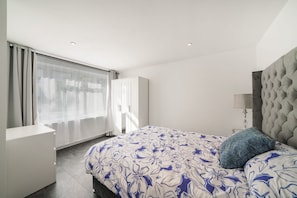 A bedroom with a bed, a wardrobe, a nightstand, a lamp, and a window with curtains. - Ground Floor Flat with Garden & Parking (London)