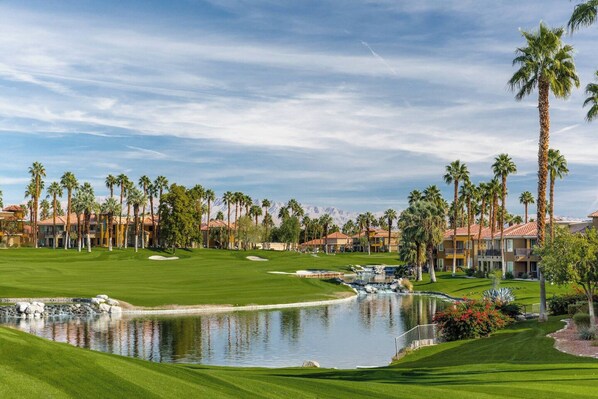 Un campo de golf con un estanque, palmeras y edificios de un resort al fondo.