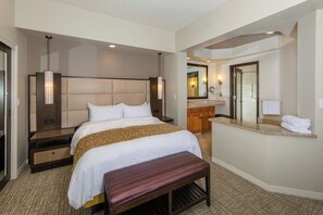 Habitación de hotel con una cama grande, una banca, un tocador con espejo y un baño con ducha y lavamanos.