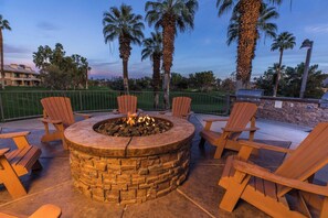 A stone fire pit with four Adirondack chairs around it, set on a patio with a view of palm trees and a golf course in the background. - Stagecoach 2027 Rental at Marriott's Desert Villas I 2 Bedroom with full kitchen (Palm Desert)