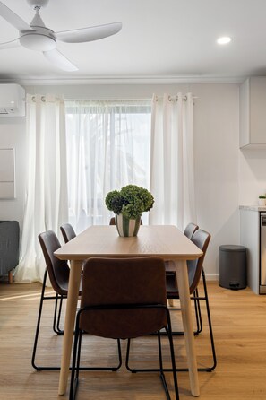 A modern dining area with a wooden table, six chairs, a ceiling fan, and a window with sheer curtains. - Onkaparinga Hideaway Beach Days and Reef Adventures (Port Noarlunga)