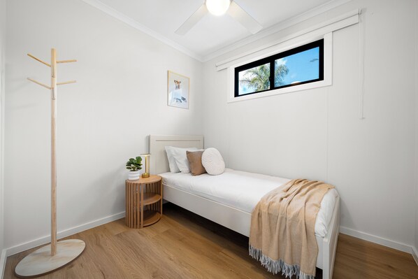 A bedroom with a single bed, a bedside table with a lamp, a coat rack, a window with a view of palm trees, and a framed picture on the wall.