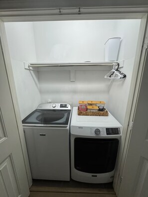 A compact laundry area with a front-loading washer and dryer, a shelf above the dryer, and a hanging rack. - Walk to Beach in 15 mins! Shared Pool/Spa! 3b/3.5b. (Port Aransas)