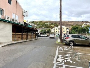 A street with parked cars, a building with a covered area, and a hill in the background. - Jamara, Central and Comfortable (Saint-Gilles les Bains)