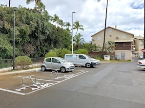 Een parkeerplaats met aangegeven parkeerplaatsen, een witte auto en een gebouw met balkons op de achtergrond.