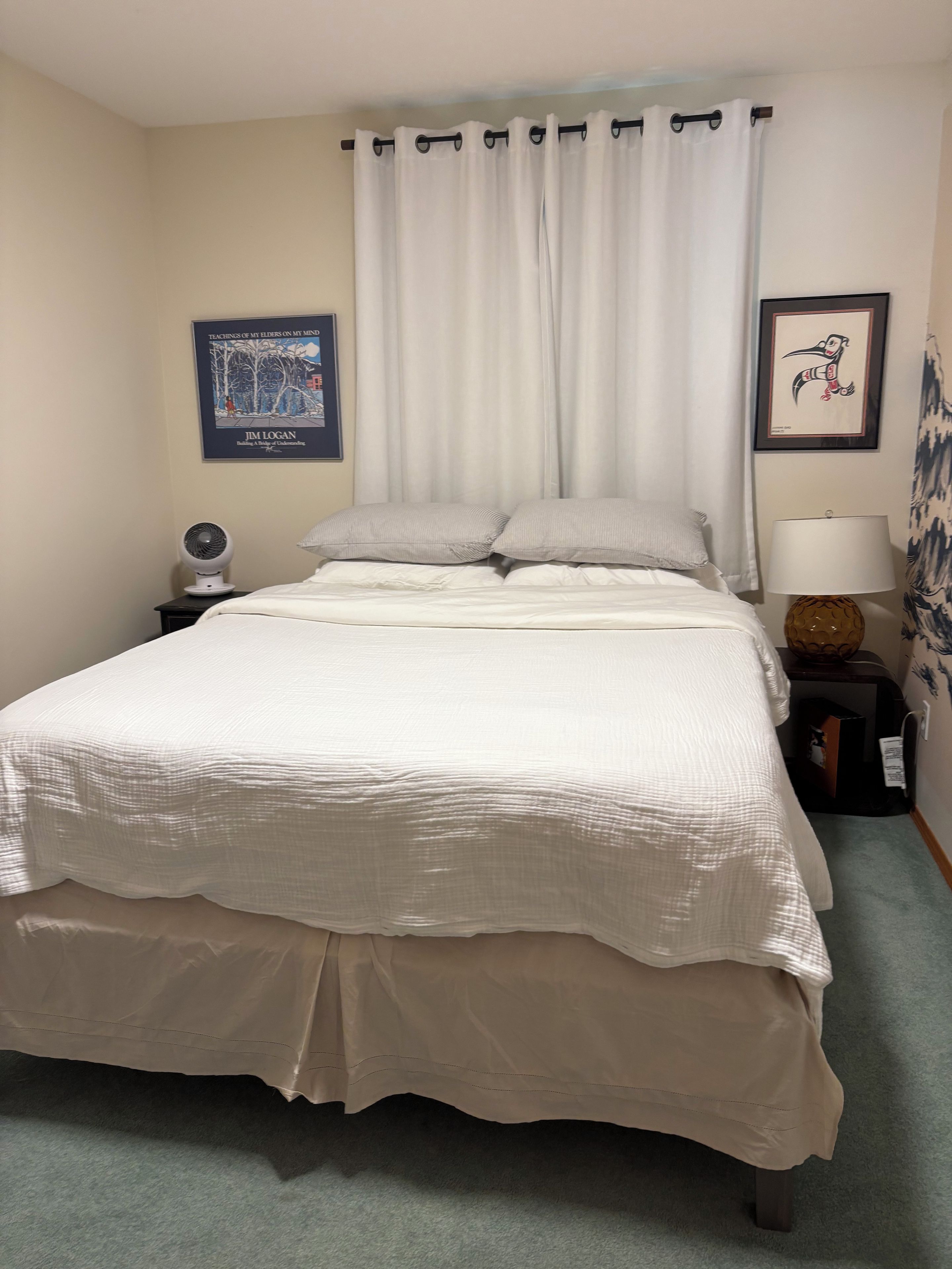 Un lit bien fait, avec une couette blanche et des oreillers, un ventilateur sur la table de chevet, un tableau encadré au mur et une lampe sur une petite table d’appoint.
