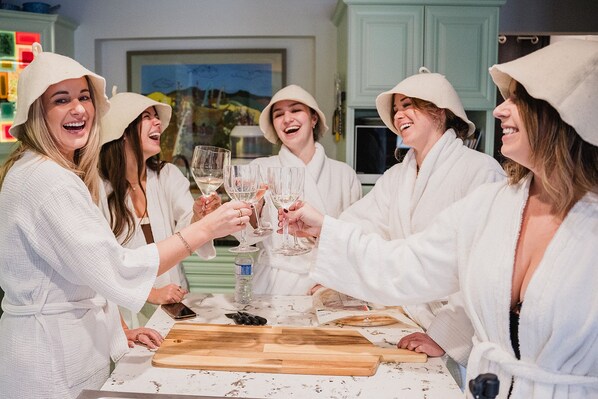 Un grupo de personas con túnicas y sombreros brindan con copas en una cocina con una tabla de cortar de madera y una botella en la encimera.