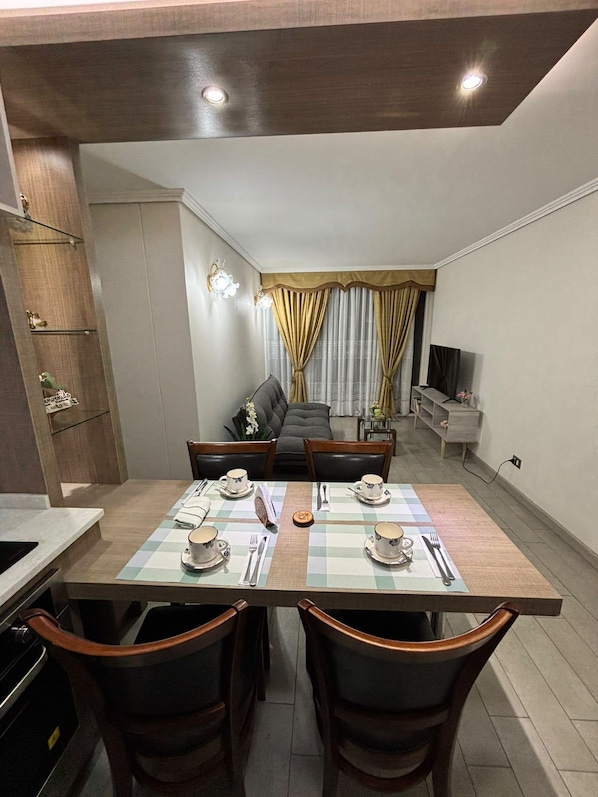 Espaço de refeições com mesa posta para quatro pessoas, sofá ao fundo e cozinha compacta à esquerda.