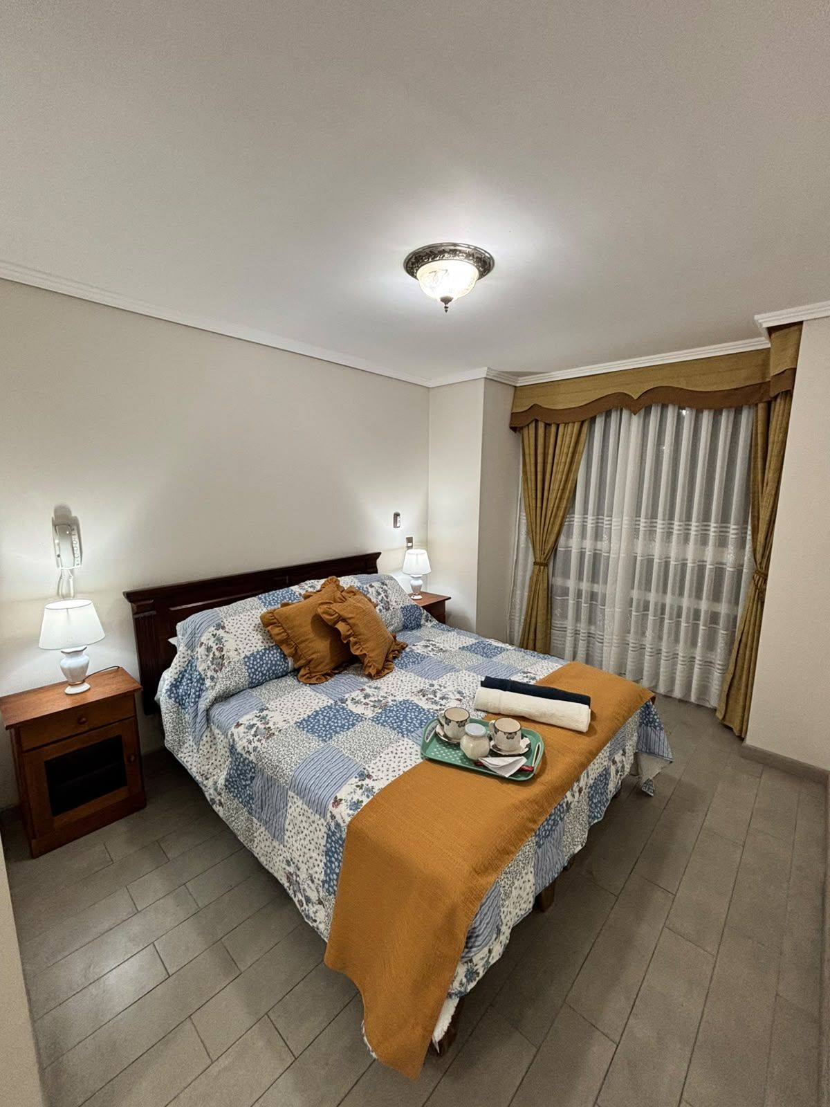 Quarto de hotel com cama, mesas de cabeceira, luminária, cortinas e bandeja com xícaras e um cobertor.