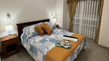 Quarto de hotel com cama, mesas de cabeceira, luminária, cortinas e bandeja com xícaras e um cobertor.