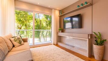 A modern living room with a sofa, a flat-screen TV mounted on a wall, a large window with a view of greenery, and a potted plant.