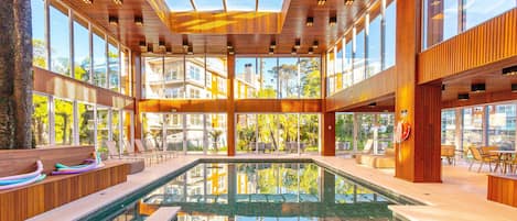 A modern indoor pool area with clear glass walls, wooden beams, and a central pool.