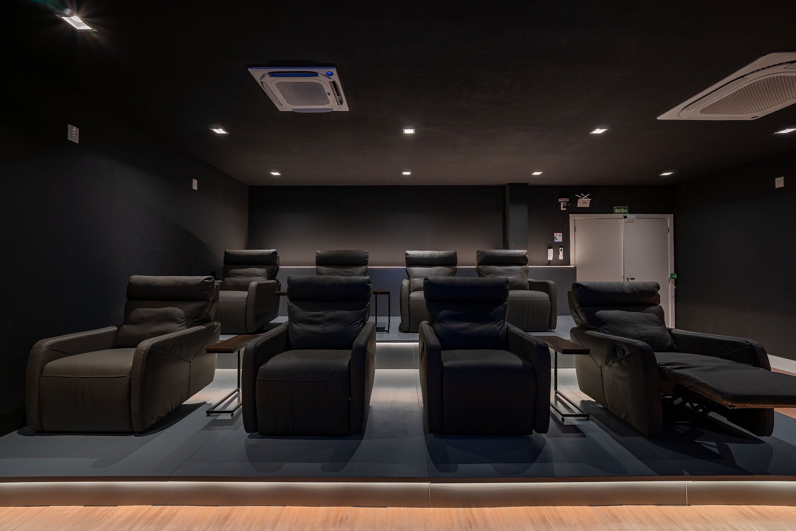 Un cine en casa con seis sillones reclinables de cuero negro, un proyector en el techo y una puerta que dice 'SAIDA'.