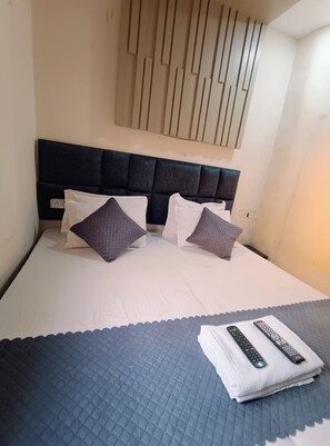 Cama com cabeceira escura, dois travesseiros, um cobertor e dois controles remotos sobre uma toalha.