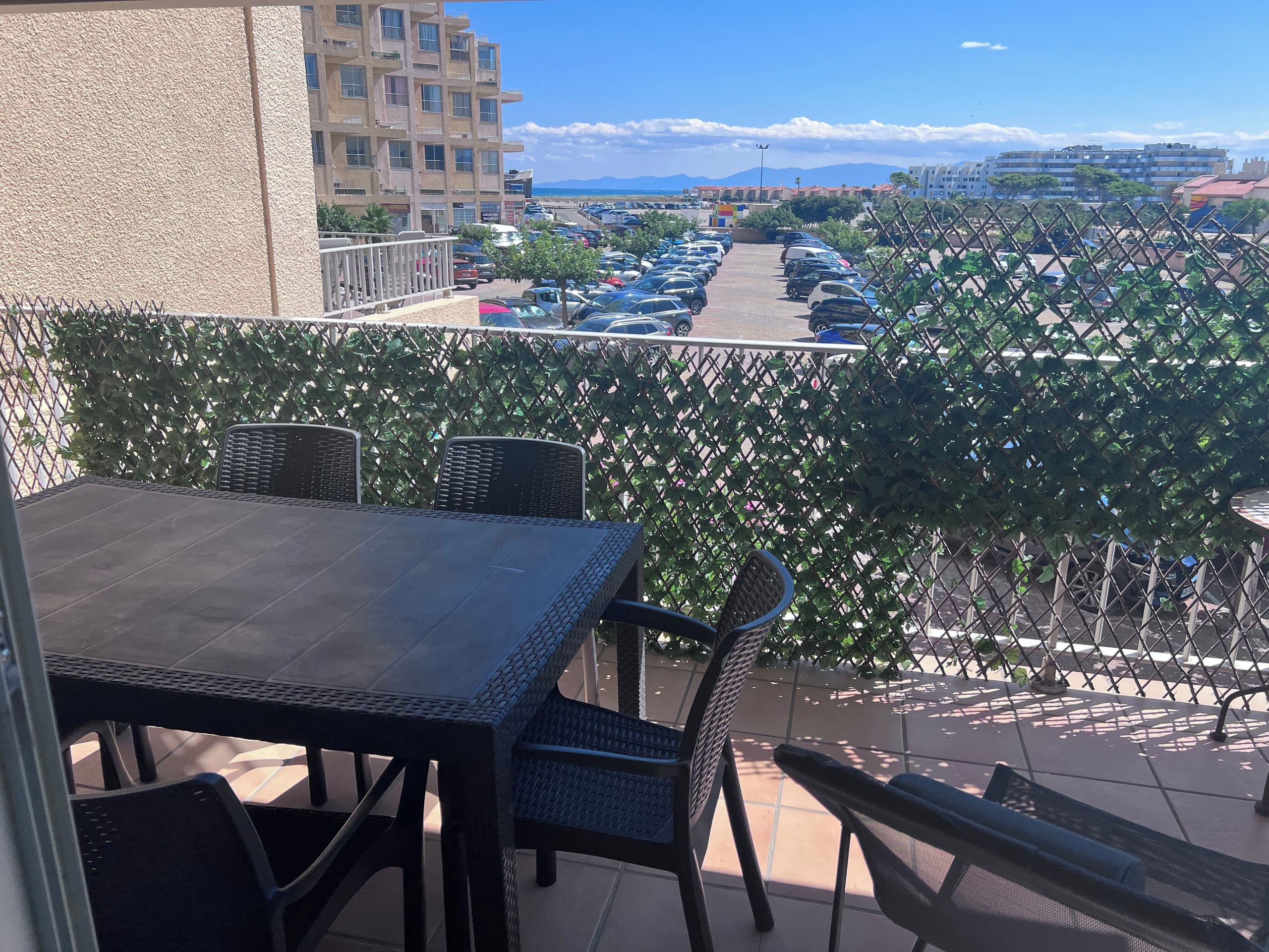 Un balcon avec une table et des chaises, donnant sur un parking et des immeubles.