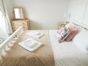 A neatly made bed with folded towels, a floral pillow, a wooden dresser, and a mirror. - Mor Awelon (Moelfre)