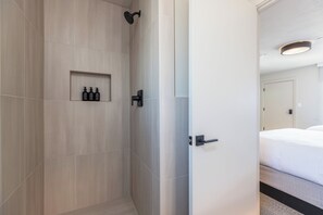 Une salle de bain d’hôtel avec une douche, une étagère contenant des articles de toilette et une porte donnant sur une chambre.