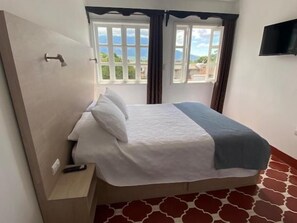 Un llit individual amb capçalera, una taula de nit i un comandament a distància a sobre. Hi ha una finestra amb cortines i un televisor fixat a la paret.