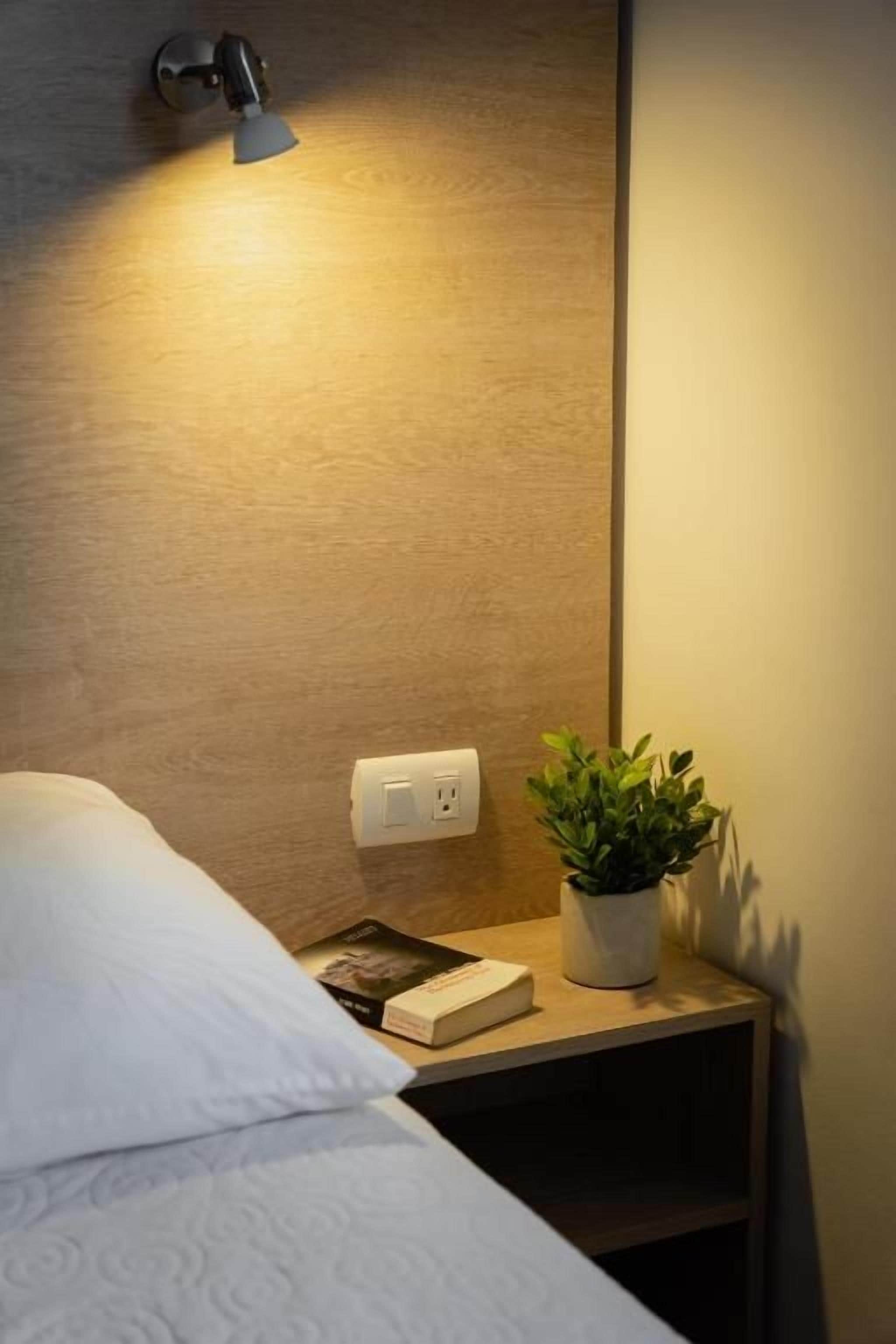 A neatly made bed with a white pillow and a book on a bedside table. There's a small potted plant on the table and a wall-mounted light fixture above.