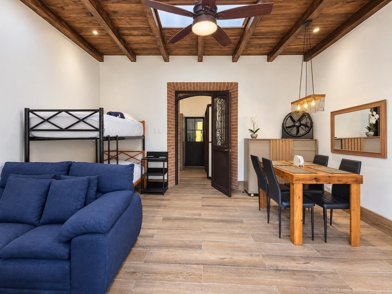 Una habitación con un sofá azul, una mesa de comedor de madera con sillas, un ventilador de techo y una pared de ladrillo.