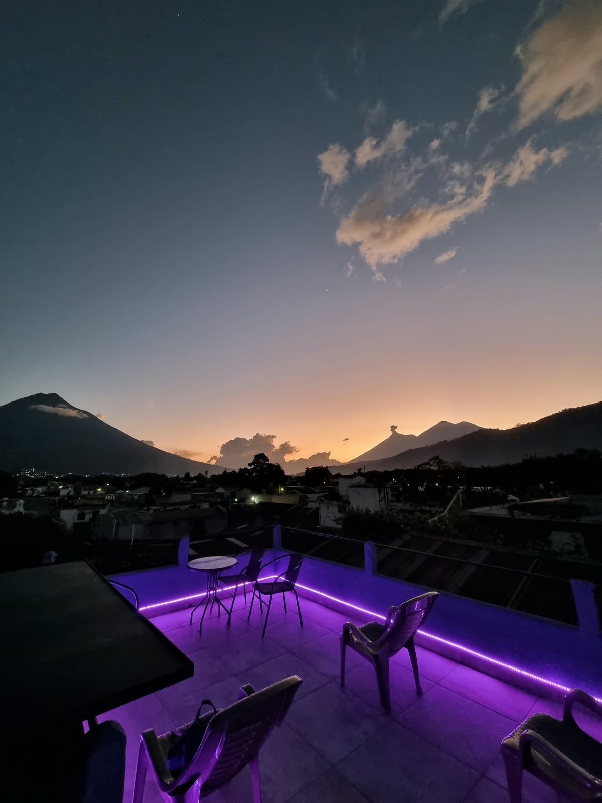 Una terraza con sillas y una mesa, con vistas a un paisaje montañoso al anochecer.