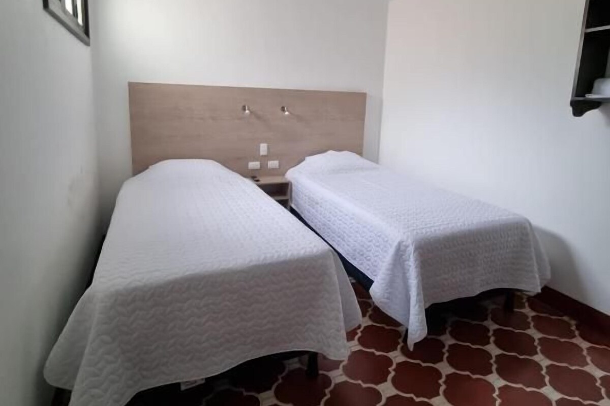 A room with two beds, a wooden headboard, a tiled floor, and a small shelf.