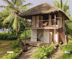 A two-story wooden house with a thatched roof, a hammock, and a chair on the balcony. - Luzul Ecohotel (Arusi)