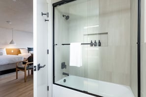 Un baño moderno con ducha envuelta en vidrio, una bañera blanca y una mesita de noche con artículos de aseo.