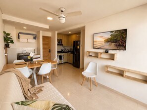 A modern living room with a sofa, dining table, chairs, a TV mounted on the wall, and a kitchen in the background. - Sienna Grove | Terrace, Caribbean Views & Pool (Cartagena)