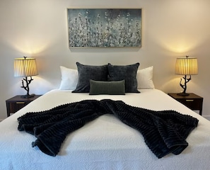 Una cama bien hecha con una colcha de diseño blanco y negro, dos lámparas de noche y un cuadro sobre la cama.