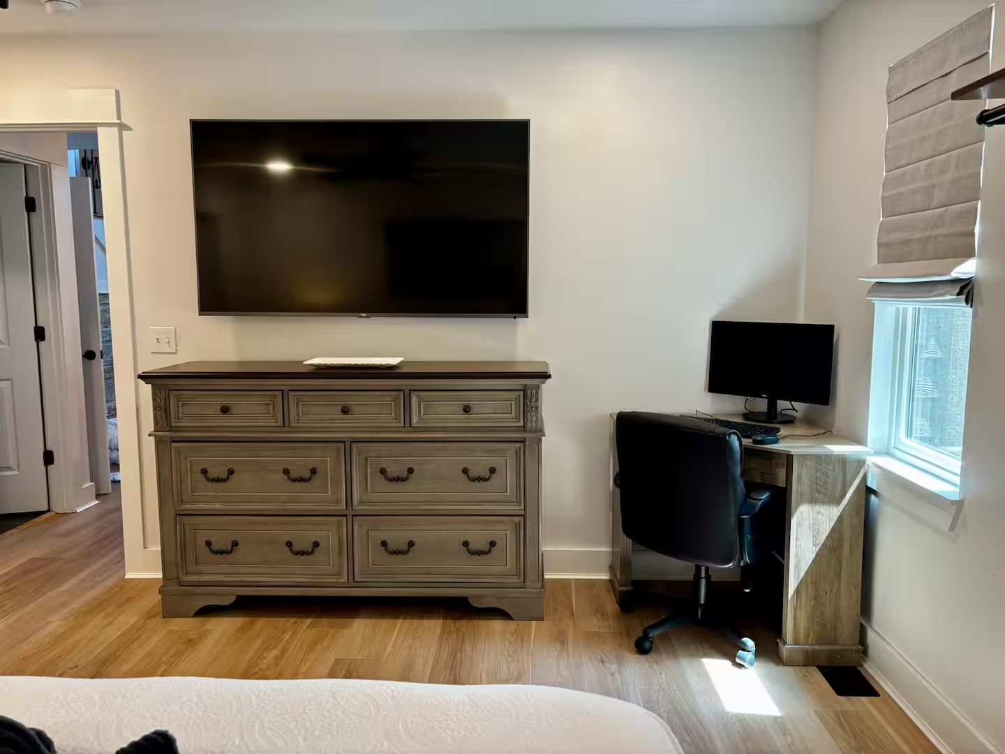 Een slaapkamer met een bed, een dressoir, een televisie, een bureau met een computer en een raam.
