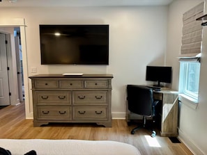 Une chambre à coucher avec un lit, une commode, un téléviseur, un bureau avec un ordinateur et une fenêtre.