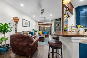 A spacious living room with a brown leather sofa, a coffee table, and a dining area with a bar. There are potted plants and a bookshelf in the background. - Indiana Jones & Jungle Cruise • Genuine Disney Décor • Secret Bookcase • Pool (Clermont)