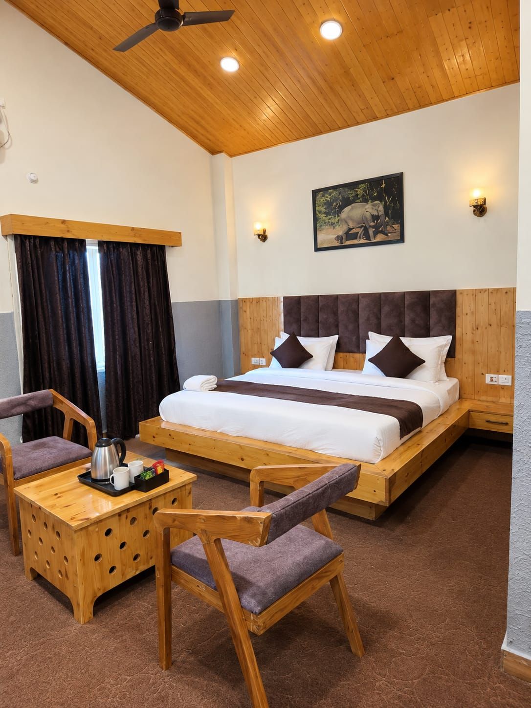 A hotel room with a wooden ceiling, a bed with a headboard, two bedside tables, a coffee table, and two chairs.