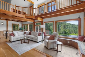 A spacious living area with a high ceiling, wooden beams, a large window overlooking a forest, and a cozy seating arrangement.