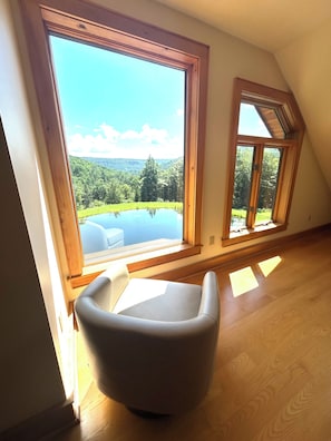 Ein Zimmer mit großem Fenster und Blick auf eine natürliche Landschaft, Holzboden und ein weißer Stuhl.