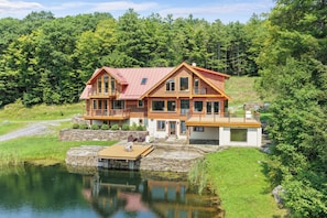 A two-story wooden house with a red roof, a stone foundation, and a dock extending into a calm lake.