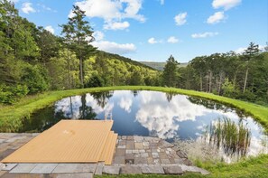 A serene outdoor setting with a wooden deck leading to a calm pond, surrounded by lush greenery and trees.