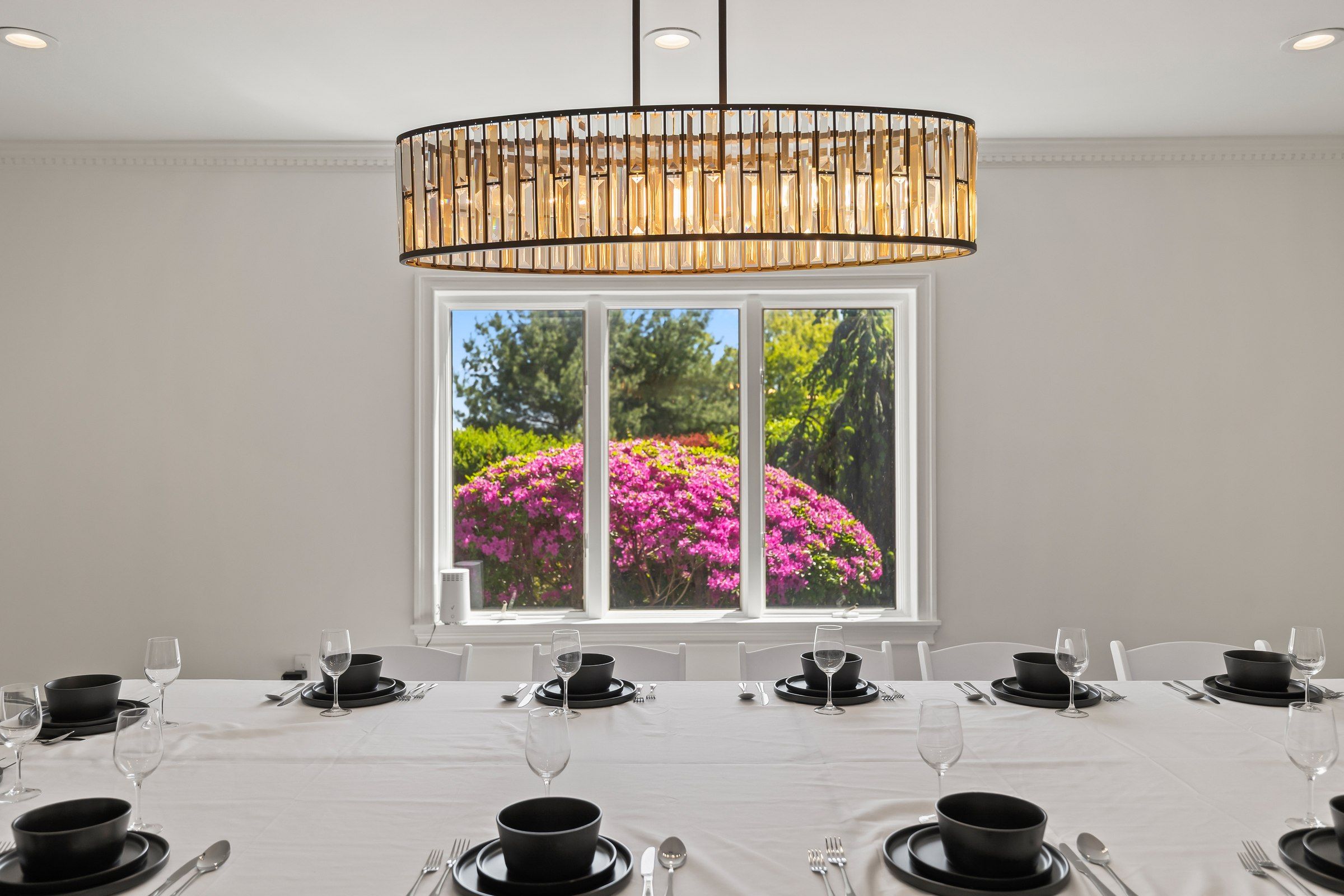 Un comedor con una mesa dispuesta para una comida, un amplio ventanal con vistas a flores rosadas y una lámpara colgante moderna.