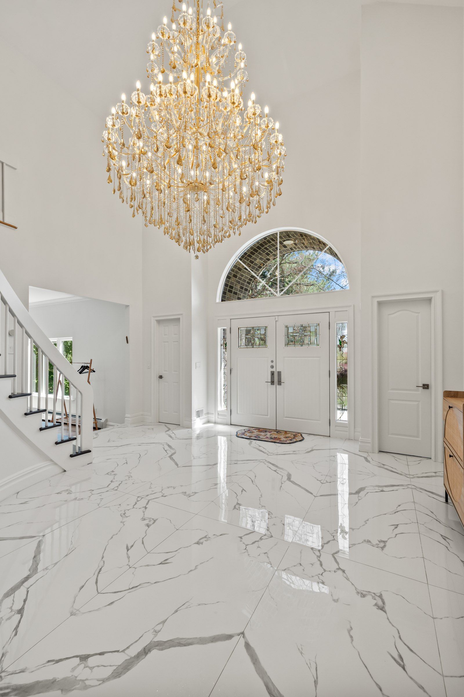 A spacious lobby with a grand chandelier, a staircase, and a large arched doorway.