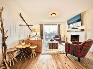 Ein Wohnzimmer mit einer Couch, einem Karoknocker, einem Couchtisch, einem Kamin und einem an der Wand montierten Fernseher.