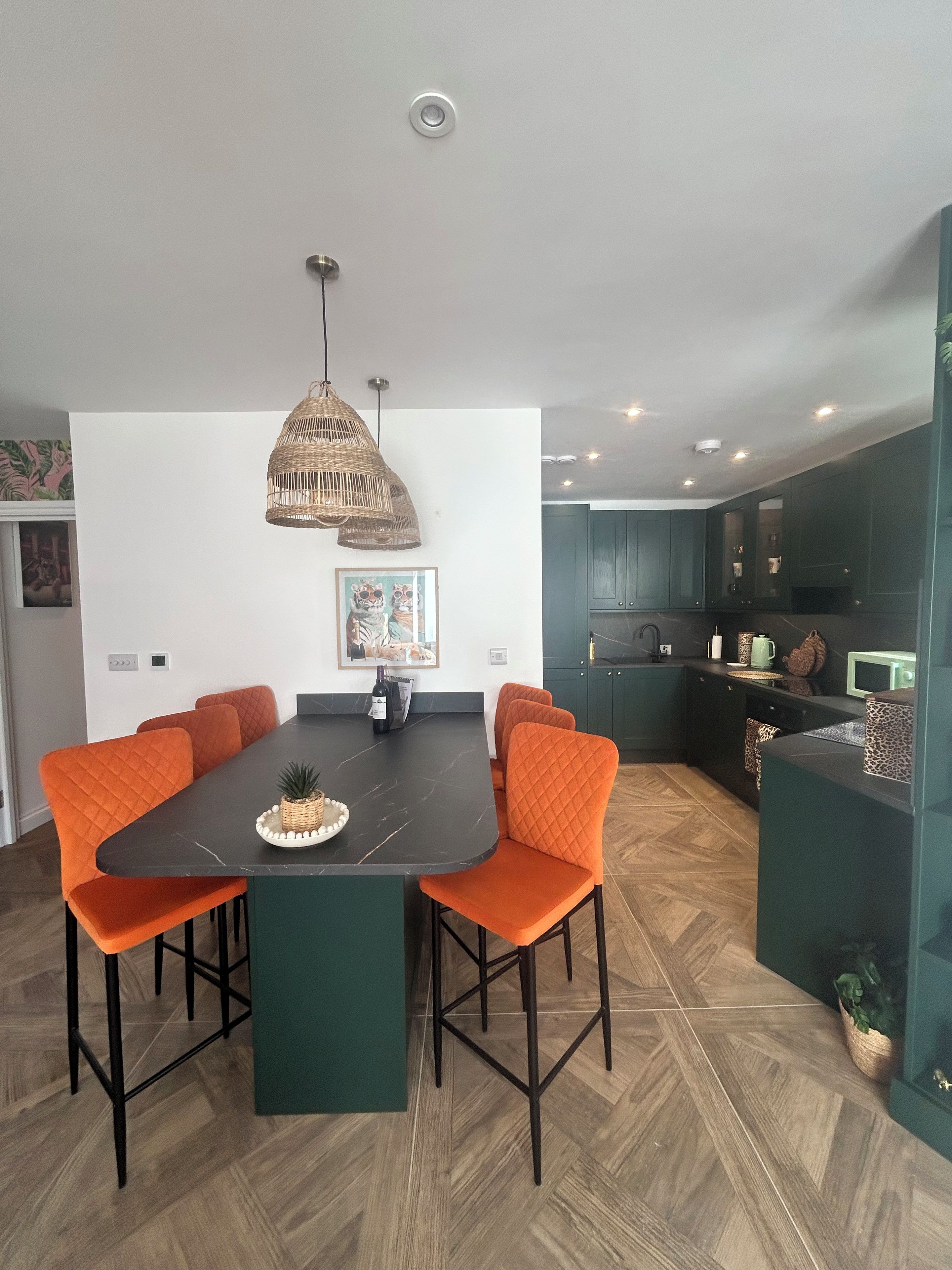 A modern kitchen with dark cabinets, a dining area with a black table and orange chairs, and a hanging pendant light.