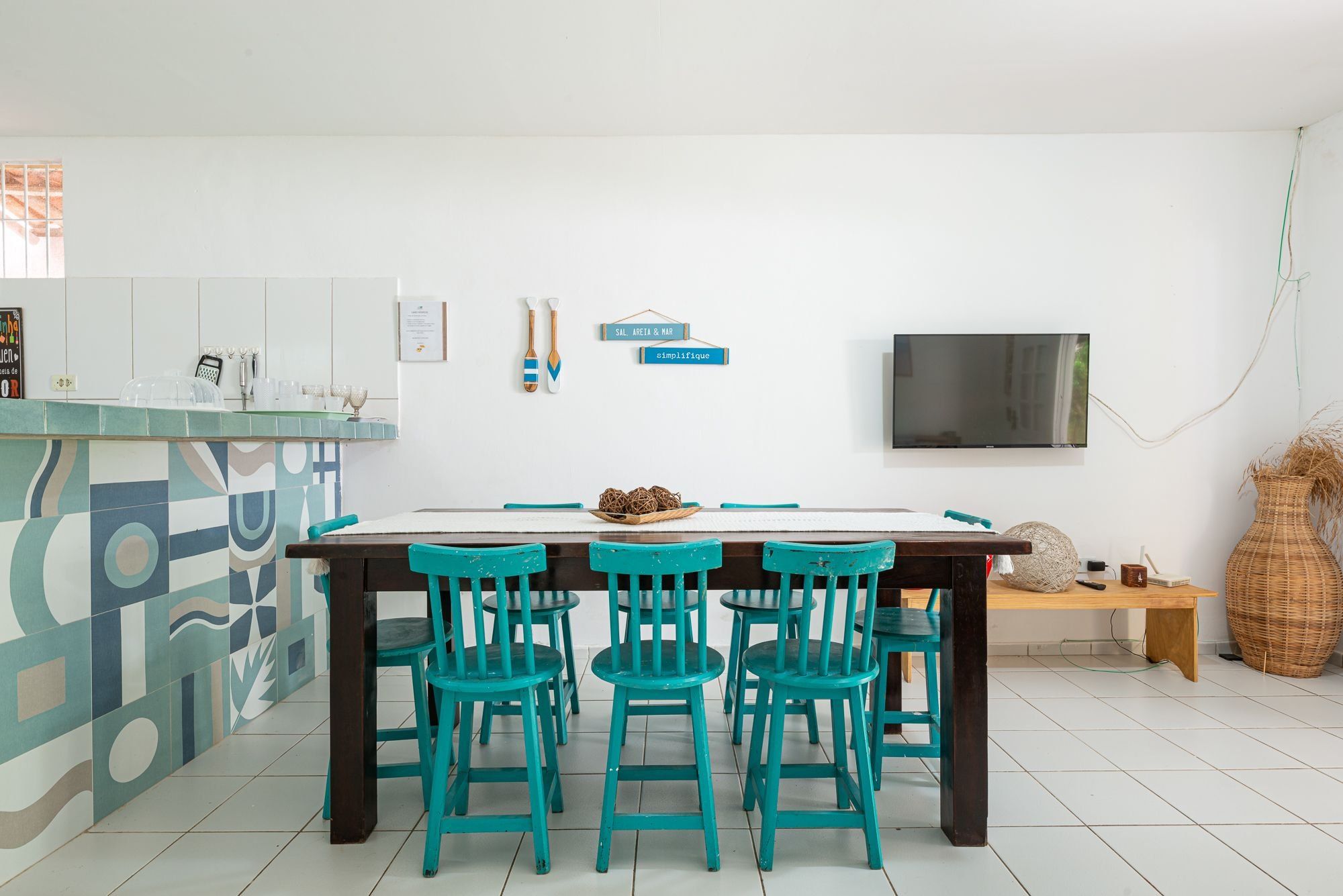 Cozinha moderna com área de refeições, piso de azulejos brancos, mesa de madeira com quatro cadeiras e televisão de tela plana na parede.
