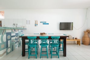 A modern kitchen with a dining area, featuring a white tiled floor, a wooden table with four chairs, and a flat-screen TV mounted on the wall.